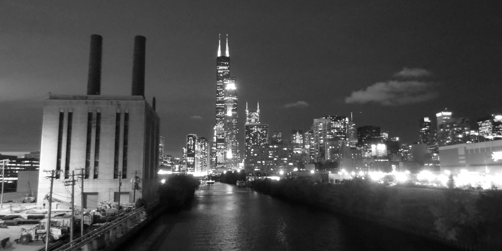 Industrial buildings and a power plant along the South Branch of the Chicago River at dusk, with the downtown Chicago skyline behind them.
