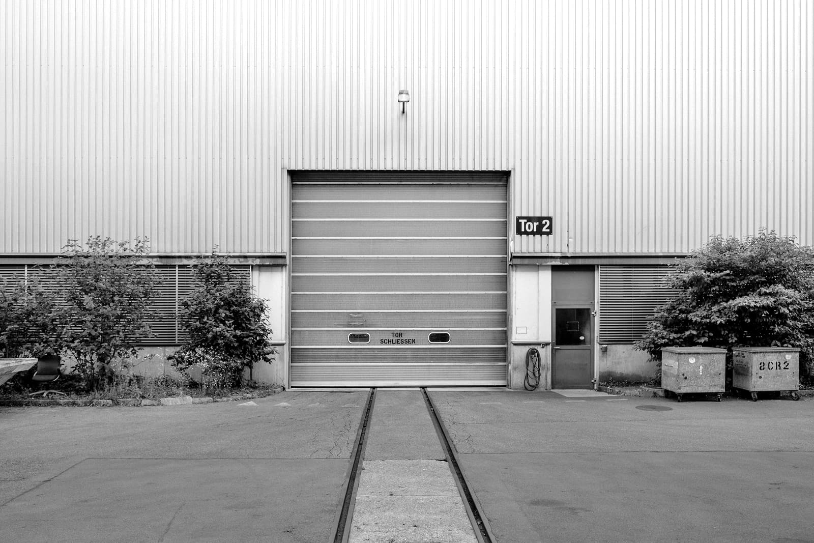 Clean corrugated-metal industrial building facade with a large roll-up door, a personnel door, and trimmed shrubs on either side.