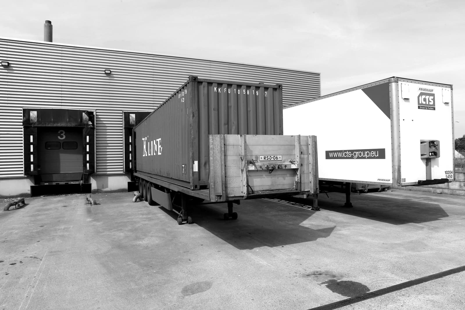 Loading dock of a warehouse with a shipping container on a chassis and a trailer backed up to dock doors.