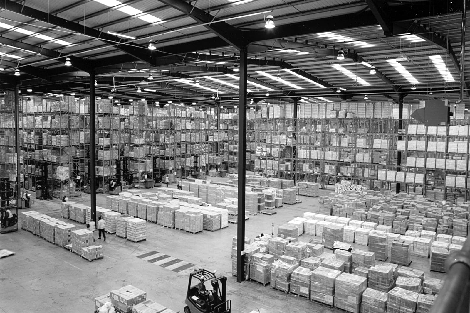 Interior of a high-bay warehouse with tall pallet racking, skylights, and forklifts moving palletized goods.